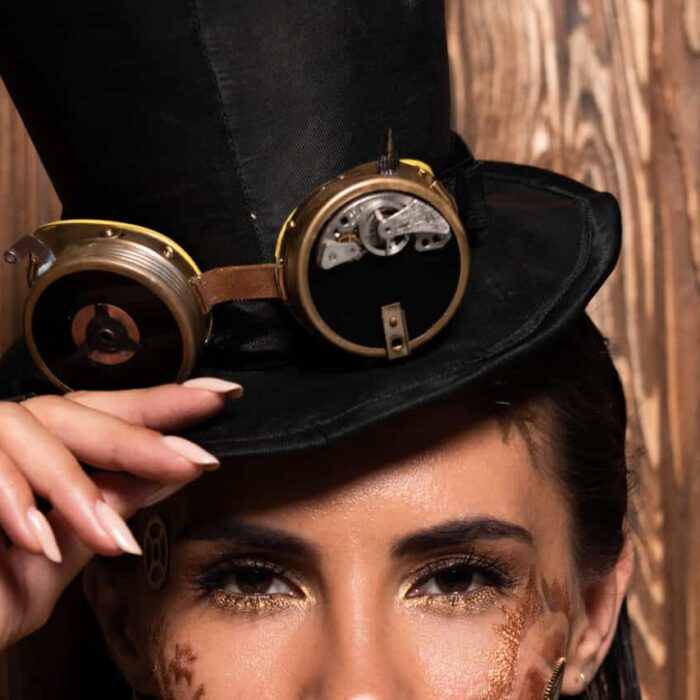 Front view of steampunk woman touching top hat with goggles looking at camera on wooden