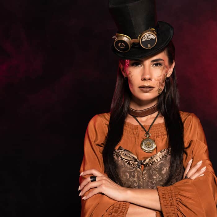 Front view of serious steampunk woman in top hat standing with folded arms on black in pink smoke