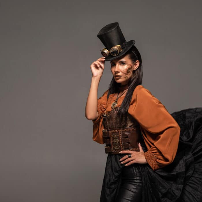 steampunk woman in top hat with goggles standing with hand on hip isolated on grey