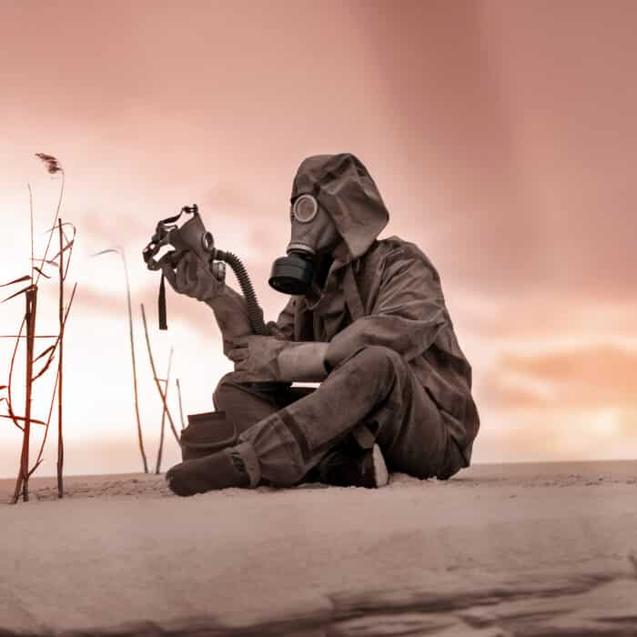 A man in a gas mask and protective suit sits on the sand and holds the gas mask of a dead friend in his hand. A man in a desperate situation. Complete loneliness. Radiation and chemical pollution.