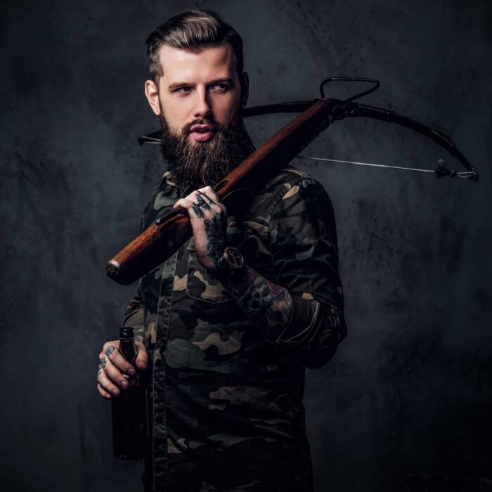 Stylish bearded hipster man in military shirt holding a craft beer and a medieval crossbow. Studio photo against a dark wall